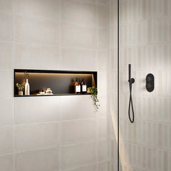 A modern shower niche with light beige rectangular wall tiles, a black backlit shelf, and black shower fixtures including a handheld shower head and control valve on a glass door.