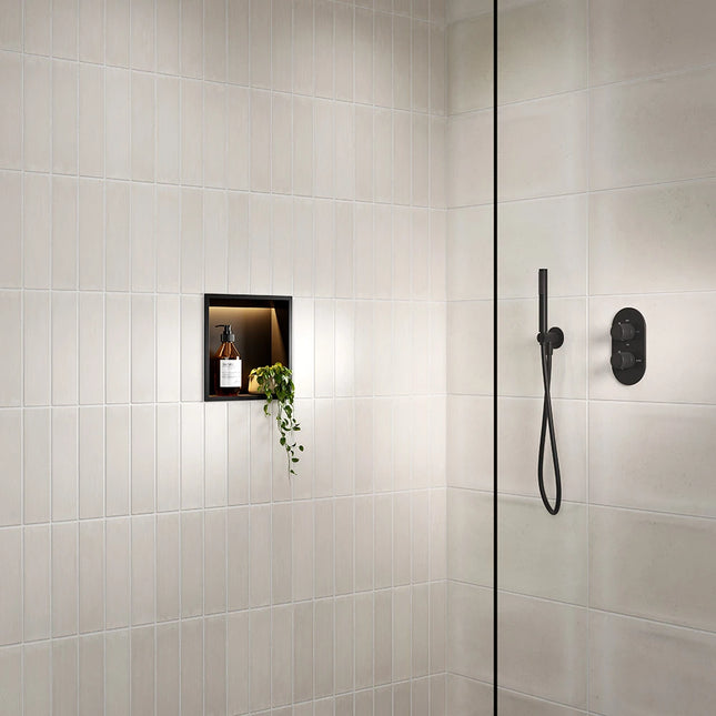 A modern shower corner with light beige rectangular wall tiles, a black-framed niche shelf holding a small bottle and trailing greenery, and matte black shower hardware including a handheld shower head and control valve.